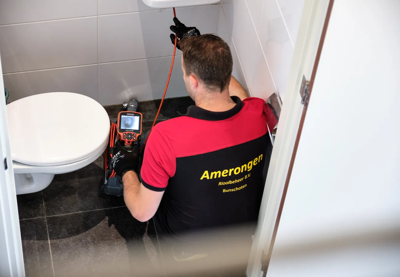 Monteur van Amerongen Rioolbeheer voert een rioolinspectie uit bij een verstopt toilet, met inspectiecamera en kabel achter de wc-pot